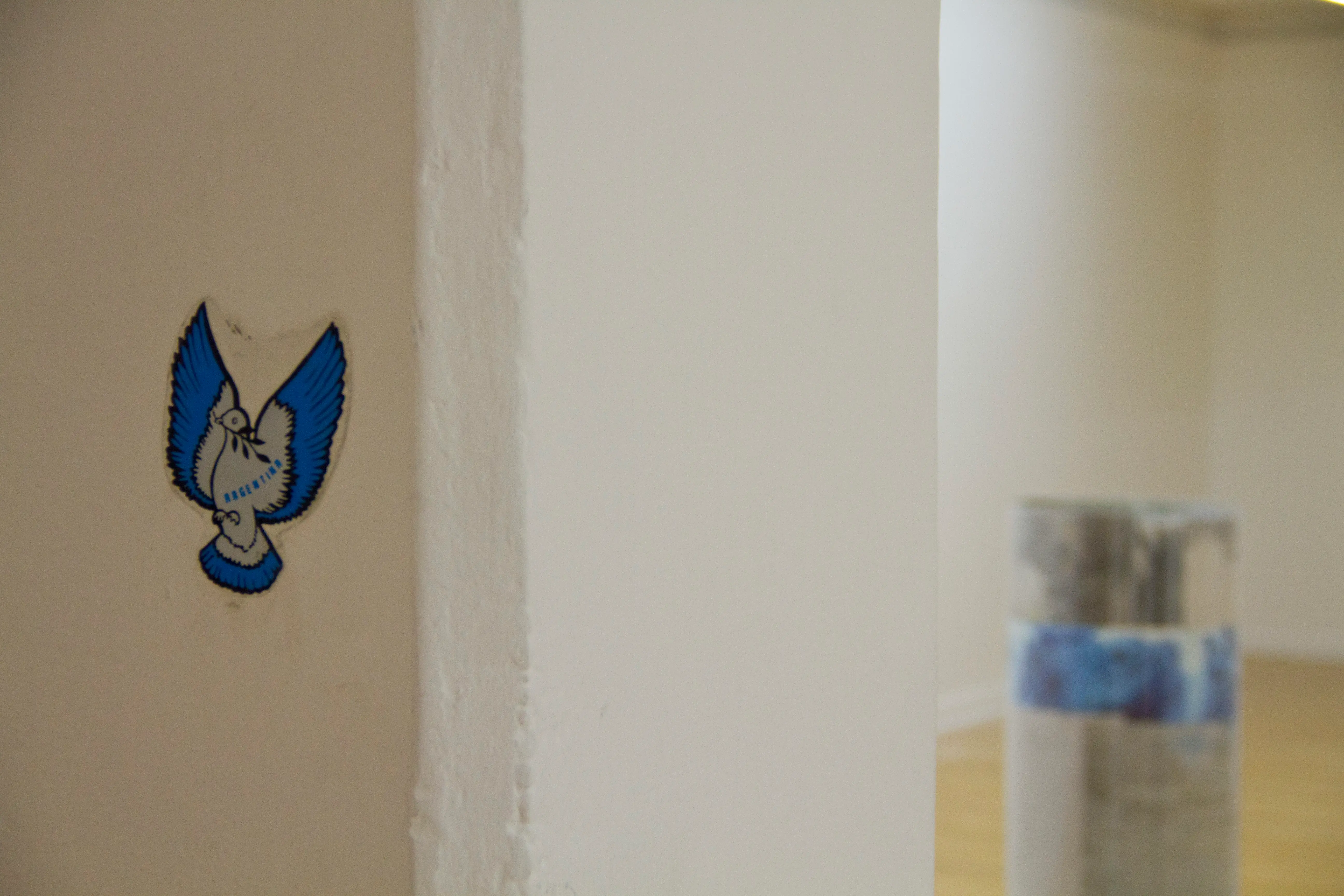 Detail from the installation, a gray dove sticker saying argentina on a column, from the show, has certain similitude to the sculpture seen in the background