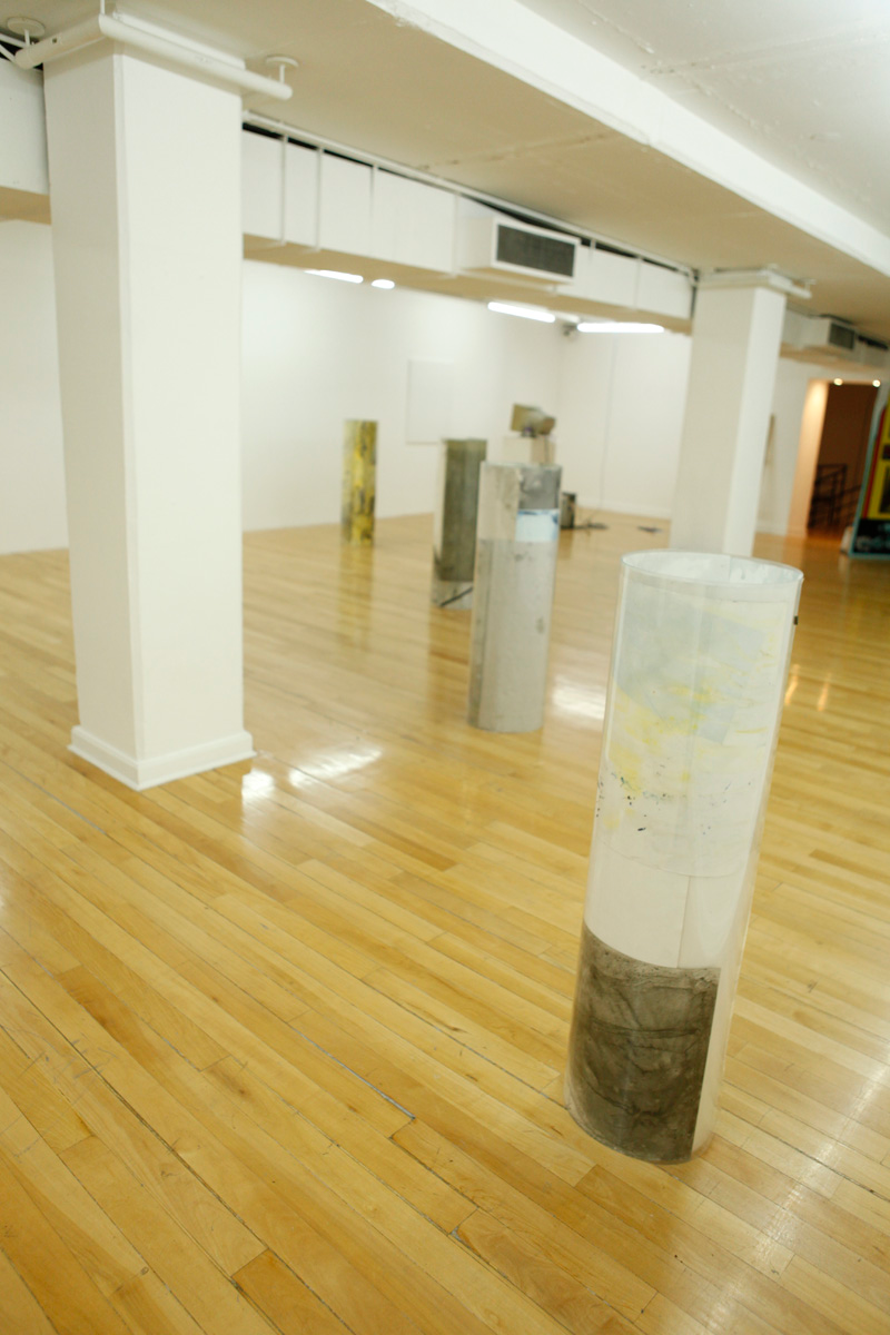 Sculpture installation where a series of drawings are rolled up between two polycarbonate sheets, creating a tube like shape standing vertically and aligned in a line, crossing tranversally the gallerys space, seen from the opposite side of the line of tube like sculptures