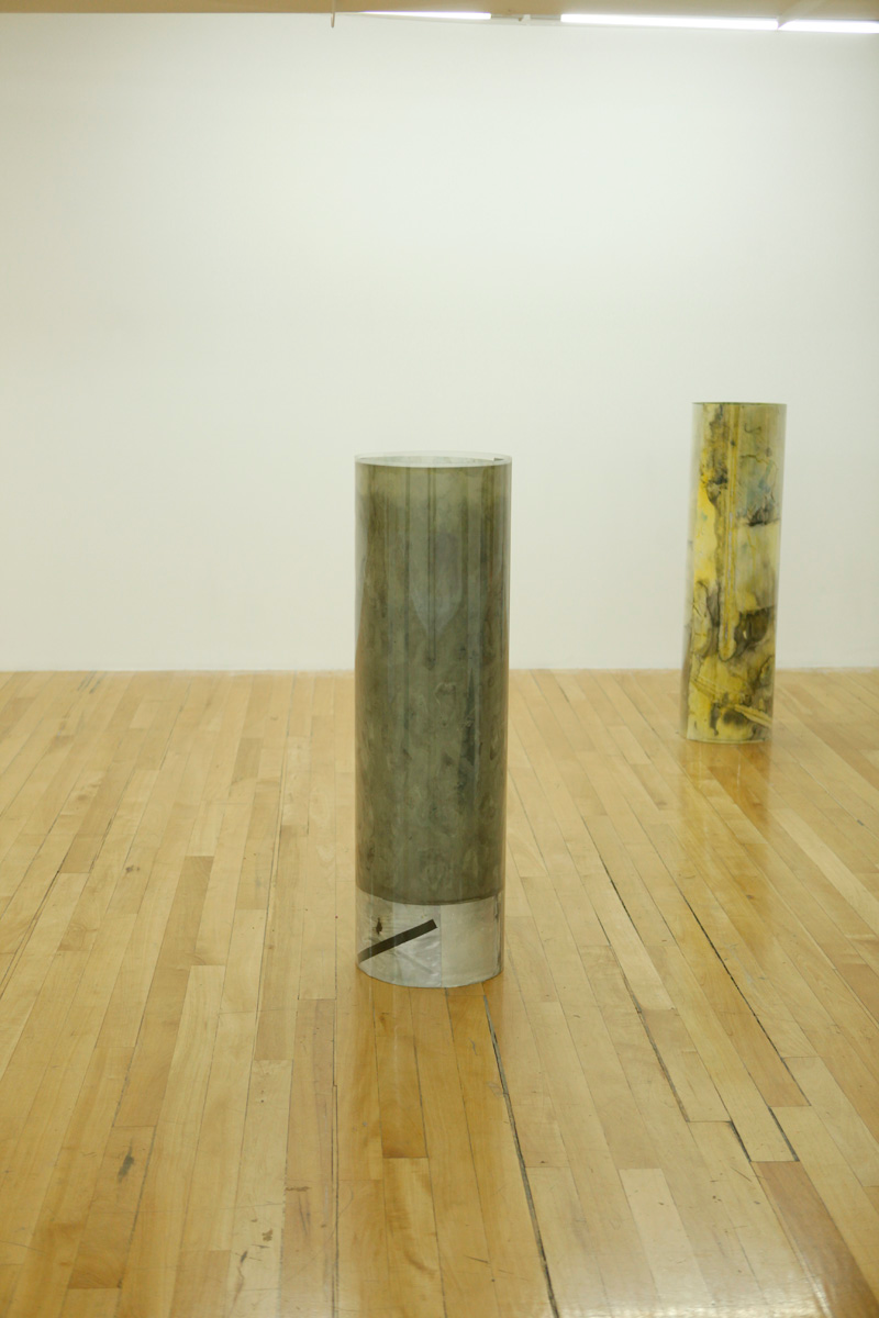 Sculpture installation where a series of drawings are rolled up between two polycarbonate sheets, creating a tube like shape standing vertically and aligned in a line, detail of two of them