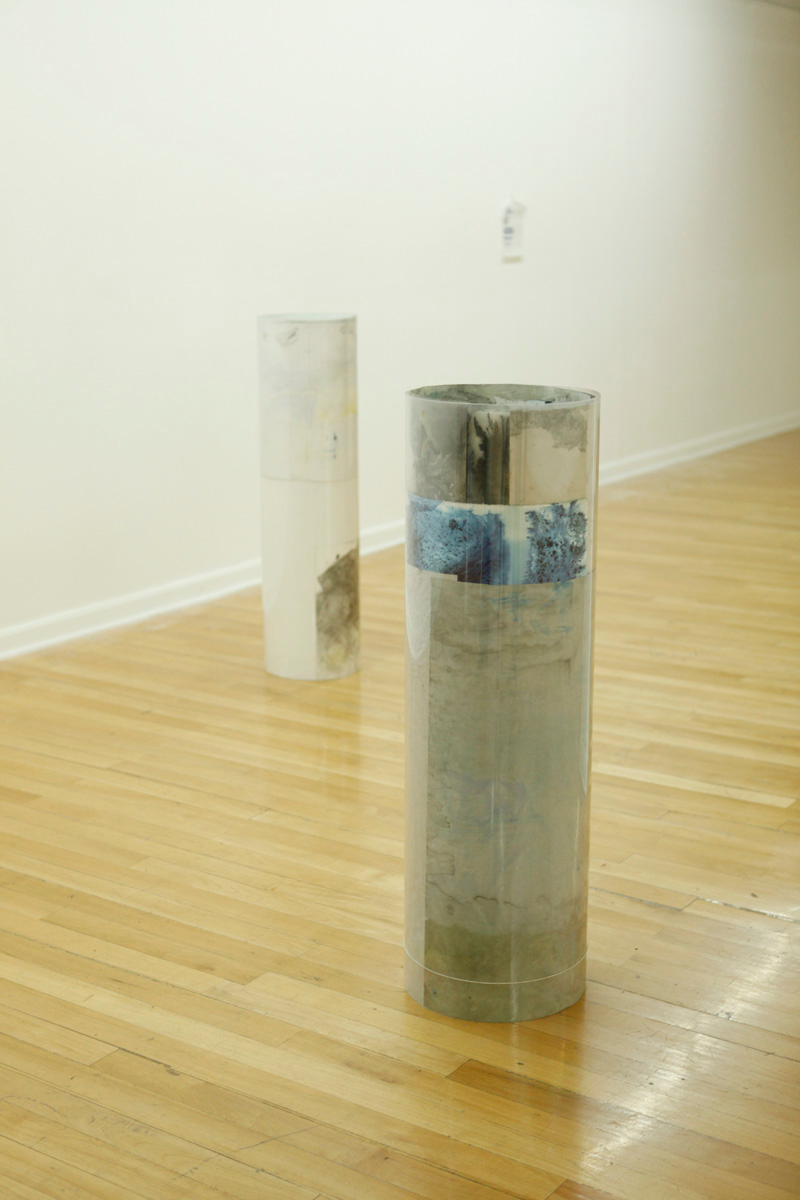 Sculpture installation where a series of drawings are rolled up between two polycarbonate sheets, creating a tube like shape standing vertically and aligned in a line, detail of two of them